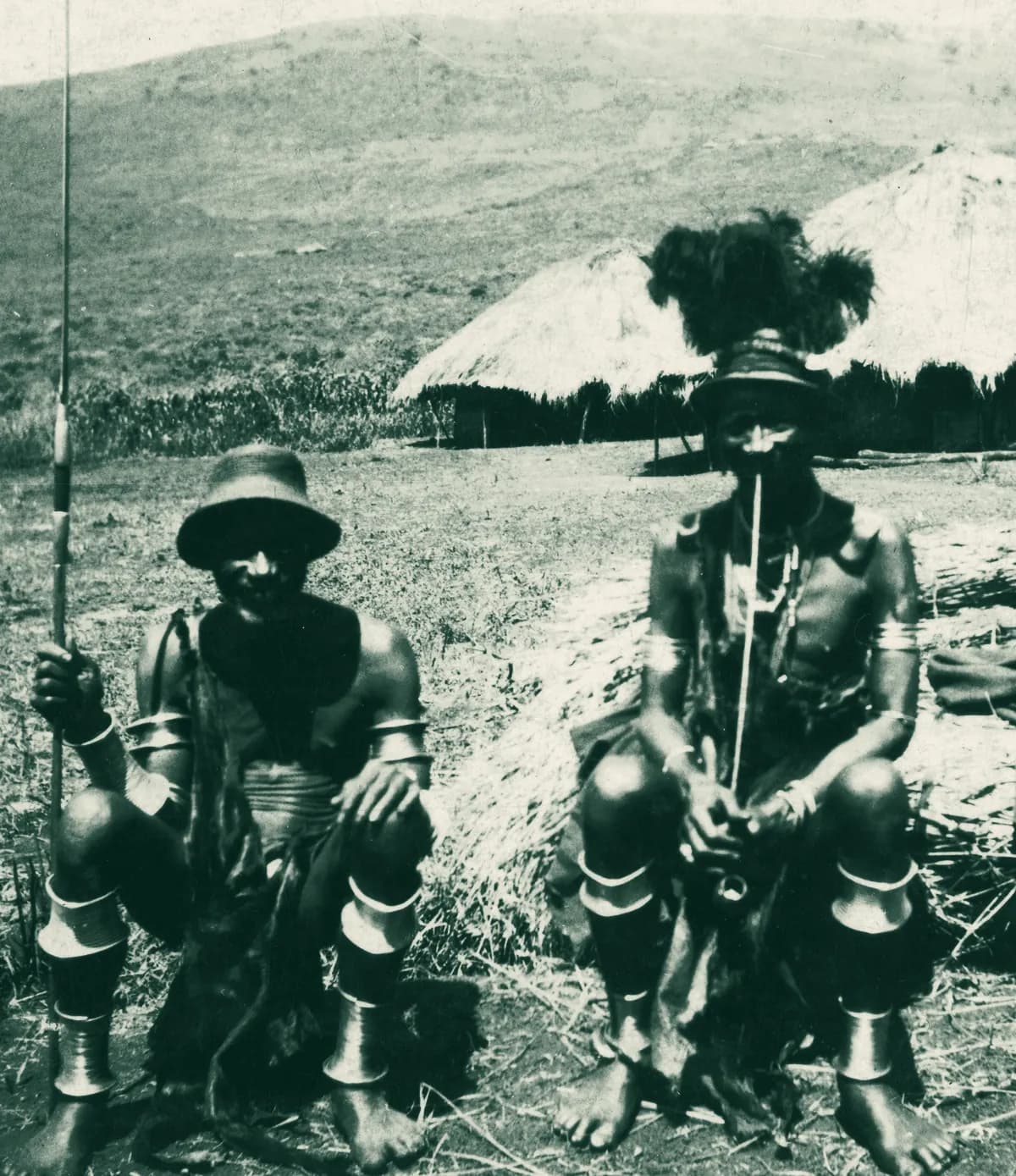 Two Abagusii elders of the Kisii native council in the 1920s, seated before thatched houses, wearing brass rings on their arms and legs.